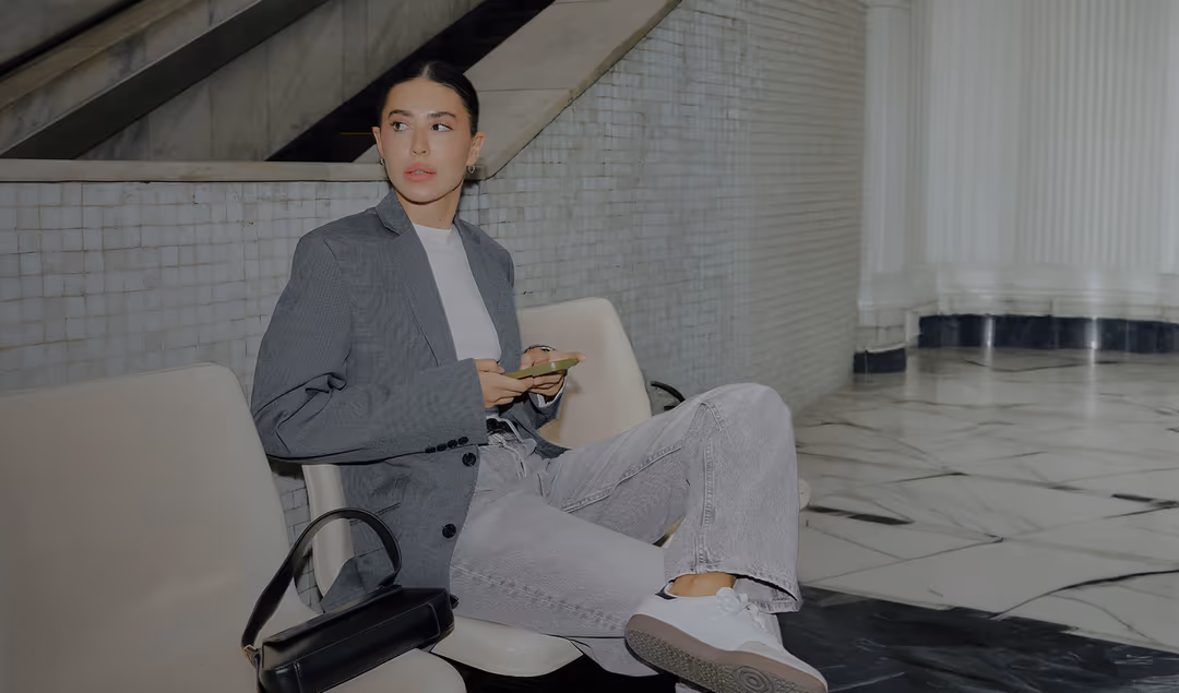 Woman wearing a gray blazer and light jeans sitting on a white chair, holding a phone, with a black handbag beside her.